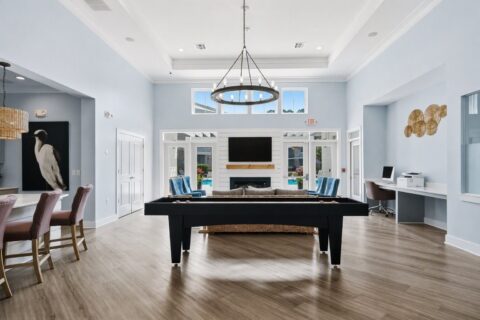 Modern clubhouse interior with light blue walls, a pool table in the center, lounge seating, a TV above a fireplace, computer desk area, and large windows letting in natural light.