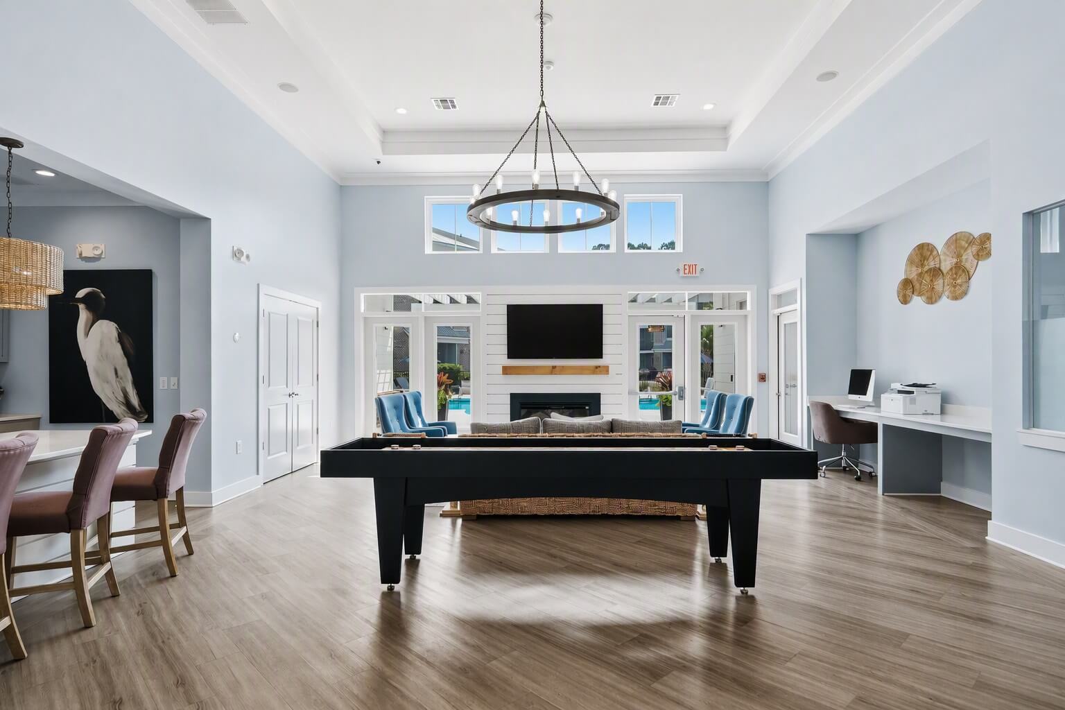 Modern clubhouse interior with light blue walls, a pool table in the center, lounge seating, a TV above a fireplace, computer desk area, and large windows letting in natural light.
