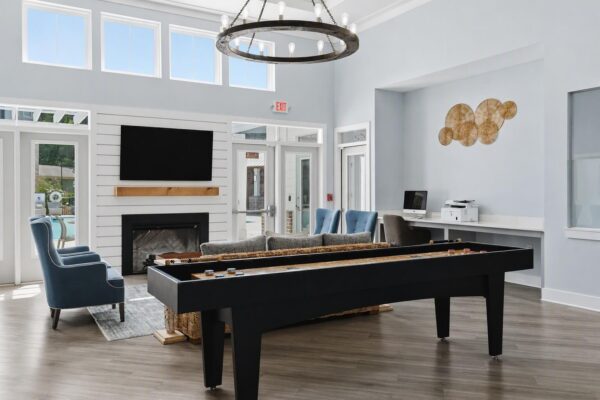 Modern lounge with shuffleboard table, blue armchairs, couch, fireplace, wall-mounted TV, computer workstation, printer, large windows, and decorative wall art. Bright, airy space with light wood floors and high ceiling.