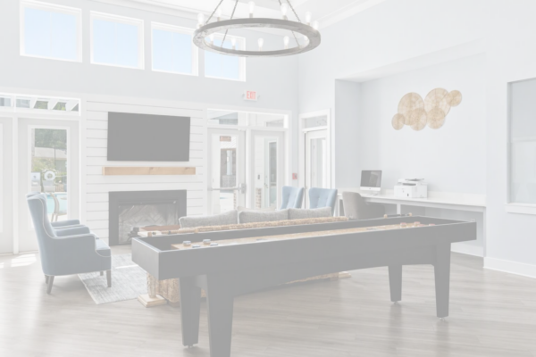 A bright lounge with large windows, a shuffleboard table, blue armchairs, a TV above a fireplace, and a desk with a computer and printer.