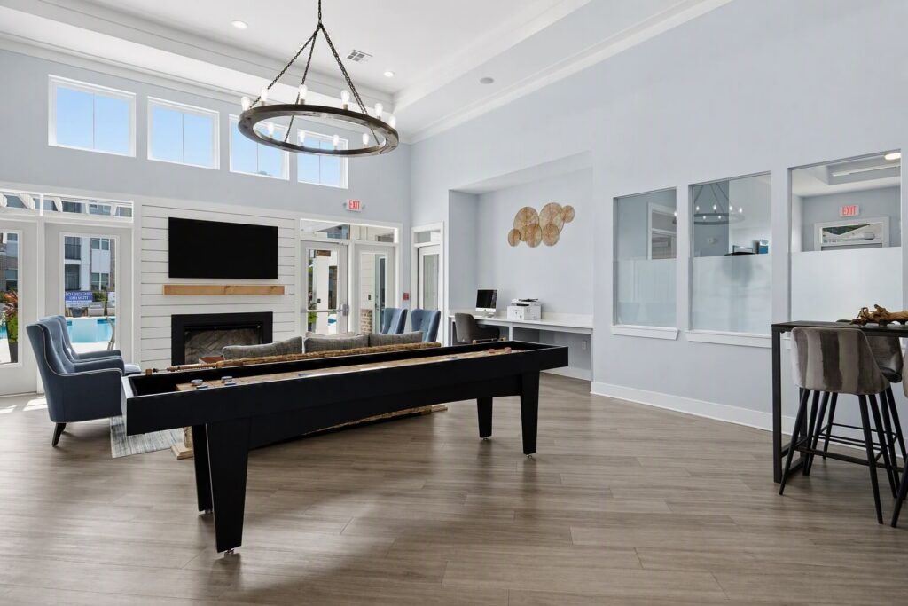 Modern lounge with a shuffleboard table, TV above a fireplace, blue armchairs, high table with stools, and large windows letting in natural light.