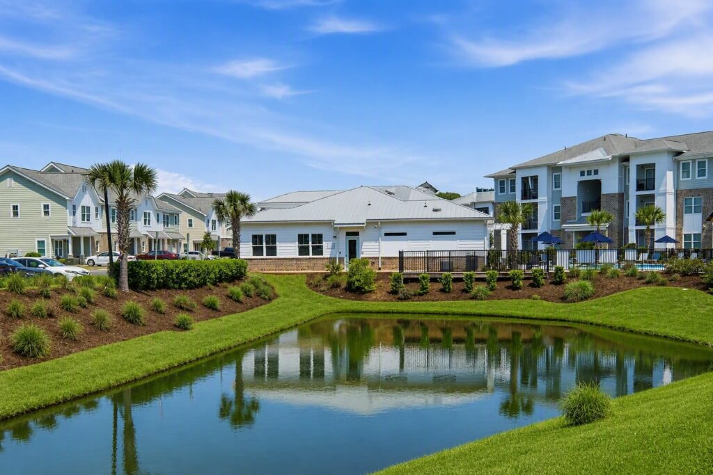 A pond surrounded by green grass, with modern apartment buildings and palm trees in the background under a blue sky with wispy clouds.