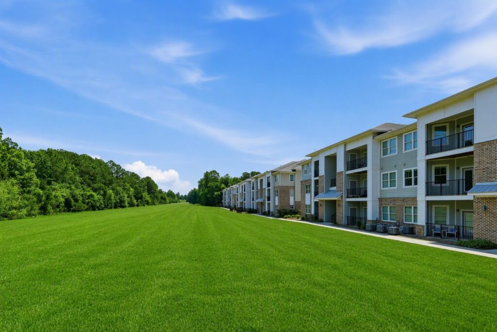 Modern apartment buildings with balconies line the right side of a large open grassy field, with trees and a blue sky in the background.