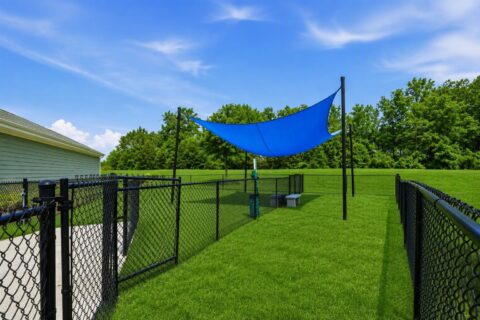 A fenced grassy area with a blue shade sail overhead, two benches, and trees in the background under a partly cloudy sky.