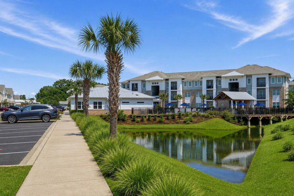 A sidewalk runs beside a pond with palm trees, leading to modern apartment buildings under a clear blue sky. Cars are parked on the left and a wooden bridge crosses the pond.