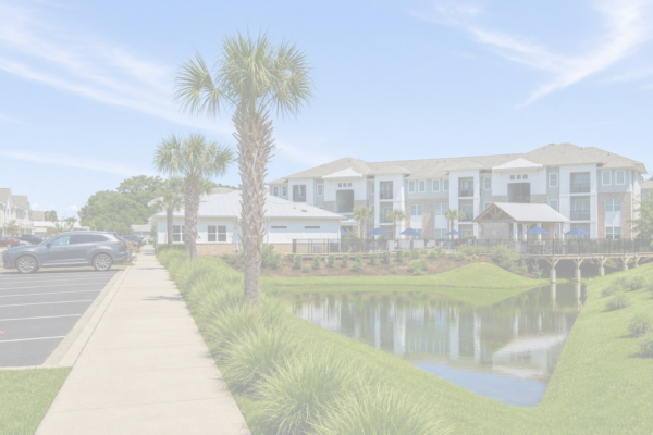 A sidewalk runs beside a pond and palm trees, leading to a modern apartment complex with balconies. Several cars are parked on the left, and a small wooden bridge crosses the pond. The sky is blue with light clouds.