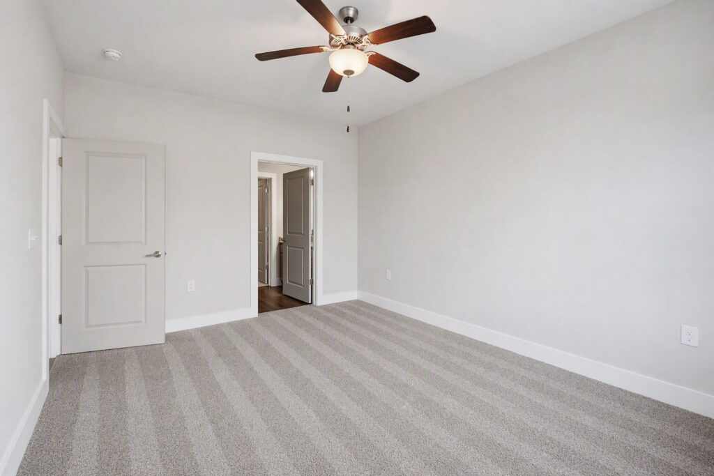 Empty bedroom with light gray walls, striped light gray carpet, a ceiling fan with lights, two closed doors, and double doors leading to a hallway with wood flooring.
