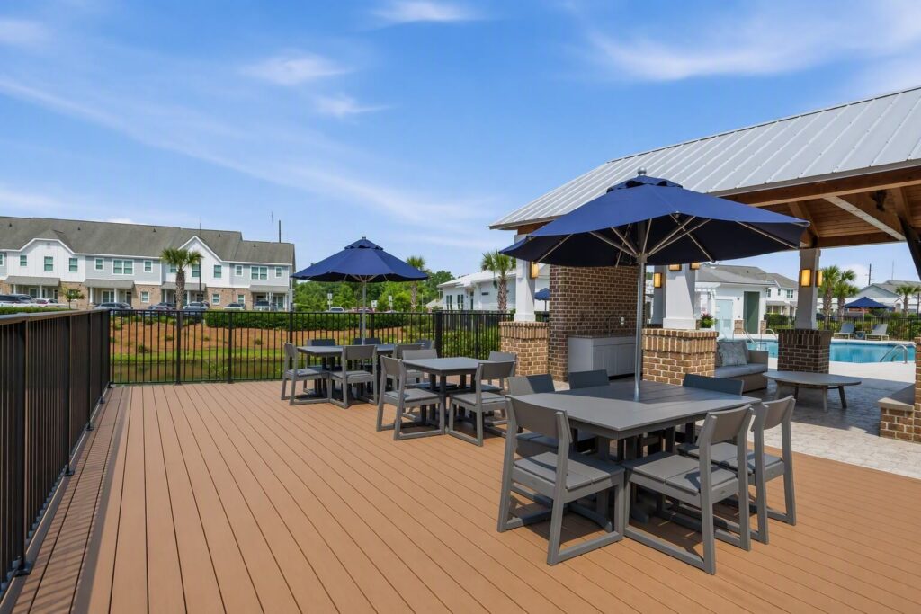 Outdoor patio with gray tables and chairs, blue umbrellas, and a covered area near a pool. Residential buildings and greenery are visible in the background under a blue sky.