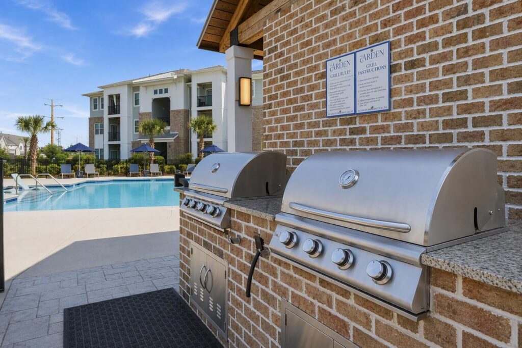 Two stainless steel grills on a brick wall near a swimming pool at an apartment complex, with lounge chairs and buildings in the background under a clear blue sky.
