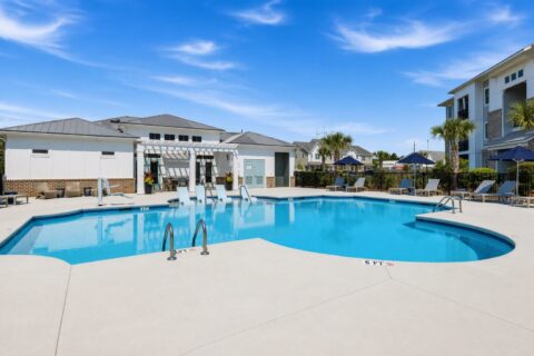 Outdoor swimming pool with clear blue water, surrounded by lounge chairs, umbrellas, palm trees, and modern apartment buildings under a bright blue sky.