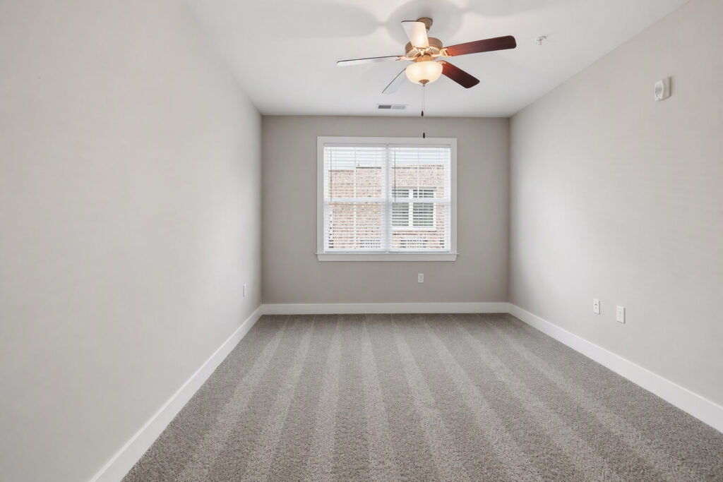 Empty room with beige walls, a gray carpet, a ceiling fan with lights, and a window with white blinds letting in natural light.