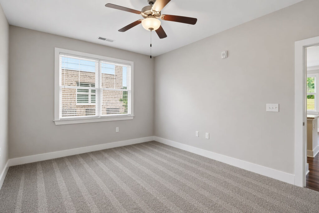 Empty room with beige walls, striped carpet, white trim, a ceiling fan with light, and a large window letting in natural light. Doorway leads to another room with hardwood flooring.