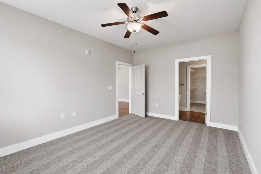 Empty bedroom with gray carpet, light gray walls, a ceiling fan with lights, an open door leading to a walk-in closet, and another door open to a hallway with wooden flooring.