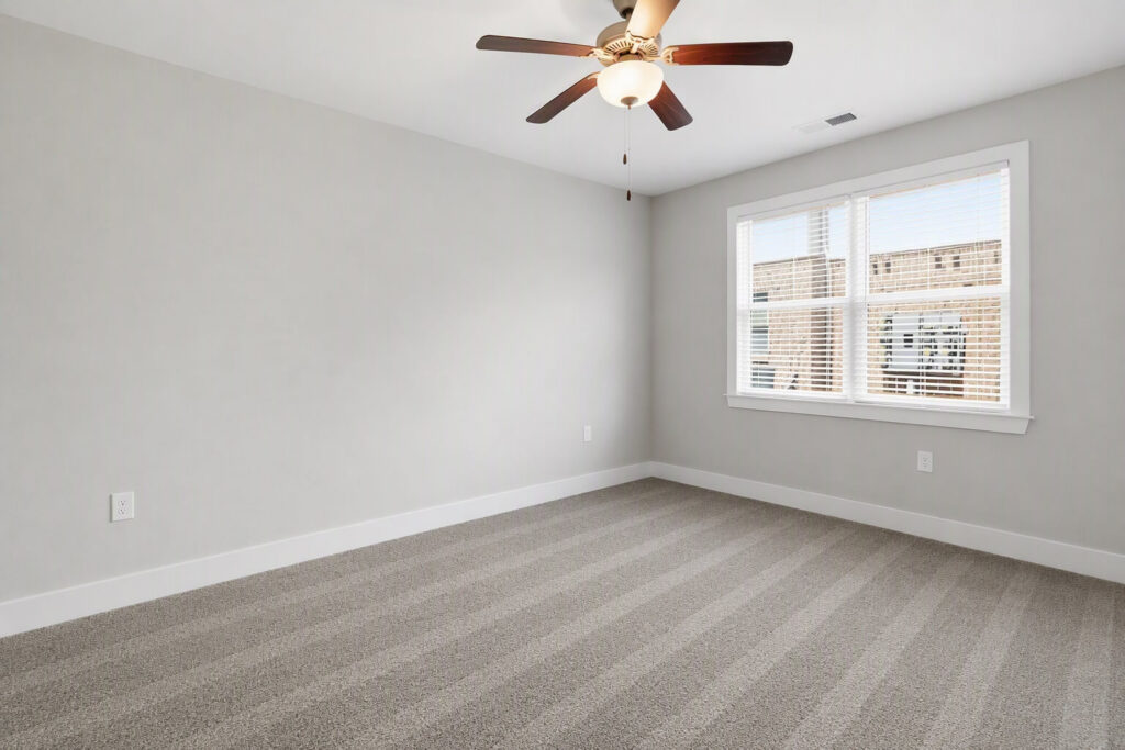 Empty room with light gray walls, striped carpet flooring, a ceiling fan with light, and a large window with blinds letting in natural light.