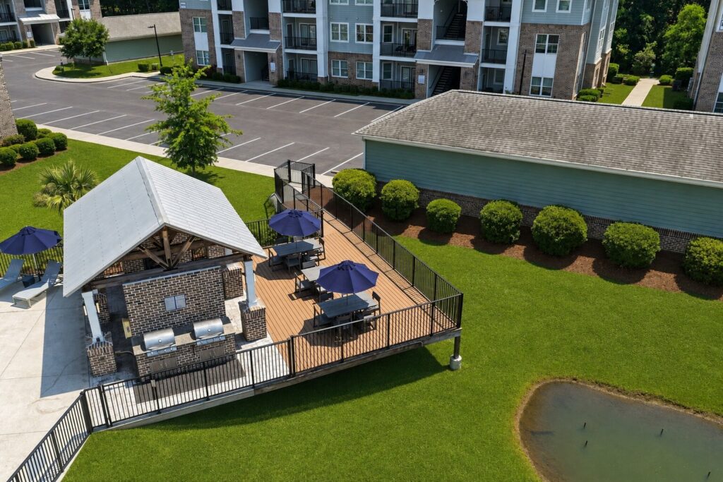 Aerial view of a small outdoor patio with picnic tables, blue umbrellas, and grills next to an apartment building, surrounded by green grass and landscaping.