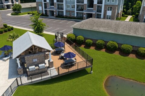 Aerial view of a small outdoor patio with picnic tables, blue umbrellas, and grills next to an apartment building, surrounded by green grass and landscaping.