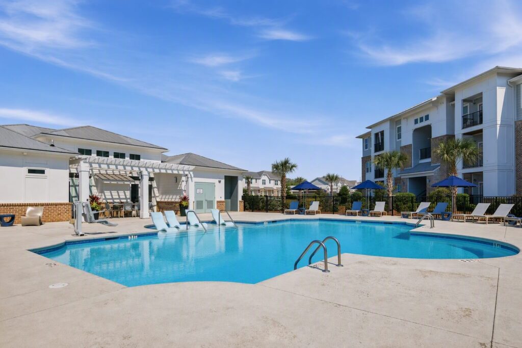 Outdoor swimming pool with lounge chairs, umbrella-shaded tables, and white buildings in the background on a clear, sunny day. Poolside seating includes in-water lounge chairs.