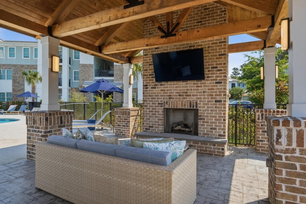 Outdoor lounge area with a wicker sofa, brick fireplace, and mounted TV under a wooden pergola. A pool, umbrellas, and modern apartments are visible in the background.