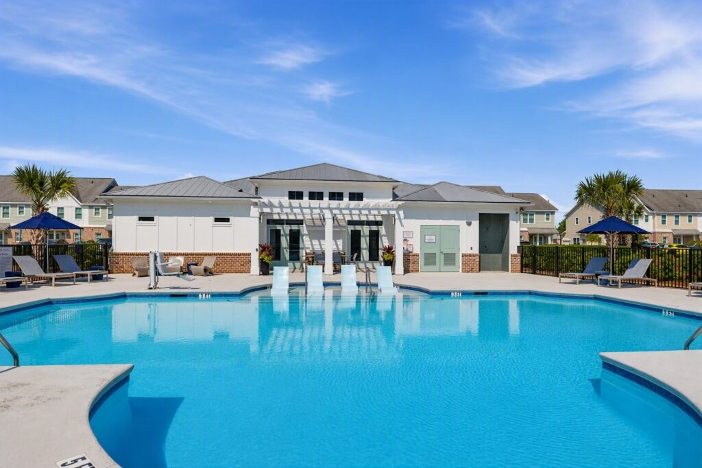 Outdoor swimming pool with lounge chairs, umbrellas, and modern clubhouse in the background under a blue sky with wispy clouds. Palm trees and townhouses are visible beyond the pool area fence.