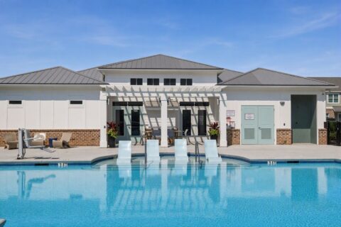 A modern outdoor pool with three submerged lounge chairs faces a building with a pergola, seating area, and clear blue sky above.