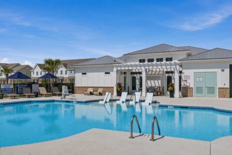 Outdoor swimming pool with submerged lounge chairs, poolside seating, umbrellas, and a modern building in the background under a clear blue sky. Apartments are visible behind the pool area.