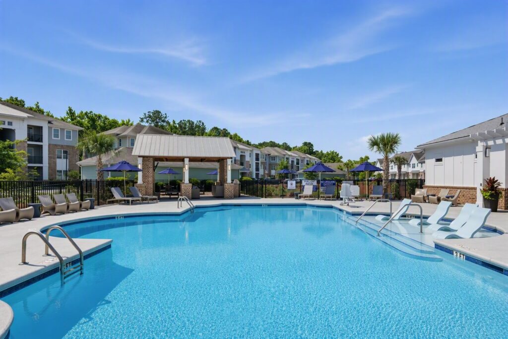 Outdoor swimming pool with lounge chairs, shaded seating areas, and umbrellas surrounded by apartment buildings and greenery under a clear blue sky.