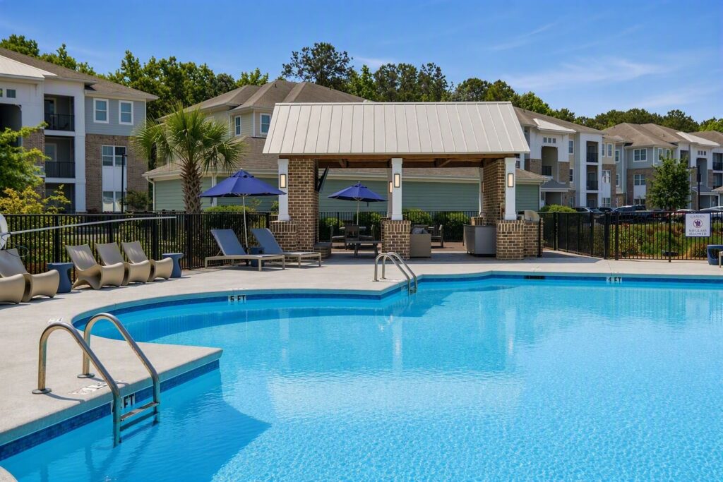 A clean outdoor swimming pool with lounge chairs, umbrellas, and a covered seating area, surrounded by modern apartment buildings and trees on a sunny day.