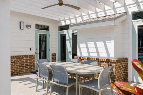 Outdoor patio with a modern dining table and six chairs under a white pergola, brick accents, and glass doors. Sunlight casts shadows on the furniture and floor. A ceiling fan is mounted above the table.