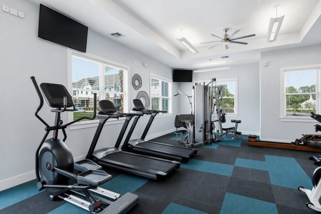 A modern gym with treadmills, an elliptical, strength machines, and large windows letting in natural light. Blue and black checkered flooring and two wall-mounted TVs are visible.