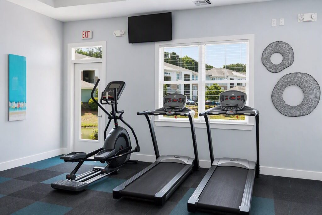 A small gym room with two treadmills and an elliptical machine, a window with blinds, wall art, and a TV mounted above the window. The floor has blue and black square tiles.
