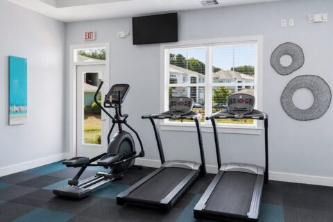 A small gym room with two treadmills and an elliptical machine, a window with blinds, wall art, and a TV mounted above the window. The floor has blue and black square tiles.