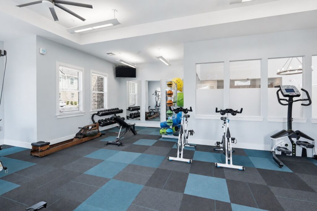 Modern gym with blue and gray checkered floor, two stationary bikes, a rowing machine, free weights, dumbbells, kettlebells, balance balls, and mirrors on the wall. Large windows provide natural light.