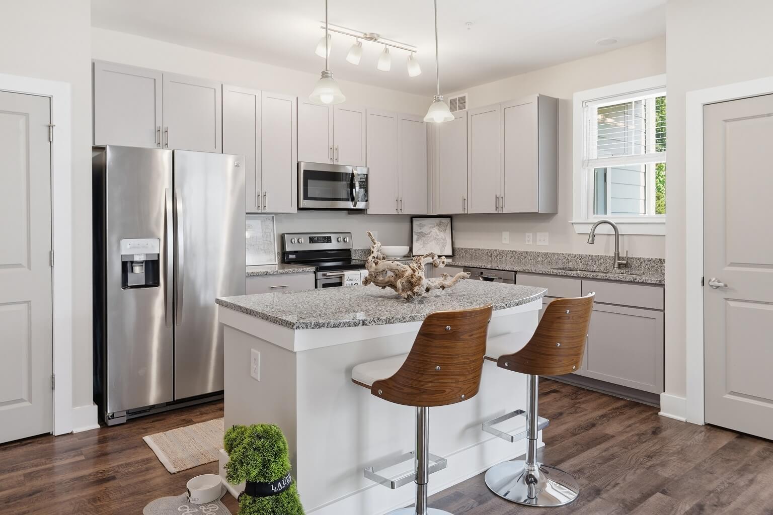 Modern kitchen with light gray cabinets, stainless steel appliances, granite countertops, island with two wooden bar stools, wood flooring, and contemporary lighting. Bright and clean space with window above the sink.