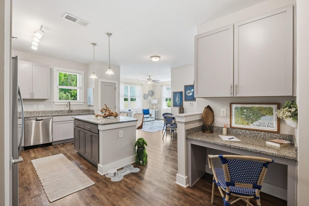 Modern kitchen with light gray cabinets, granite countertops, stainless steel appliances, a small desk area, and an open layout leading to a bright living room with large windows and wood flooring.