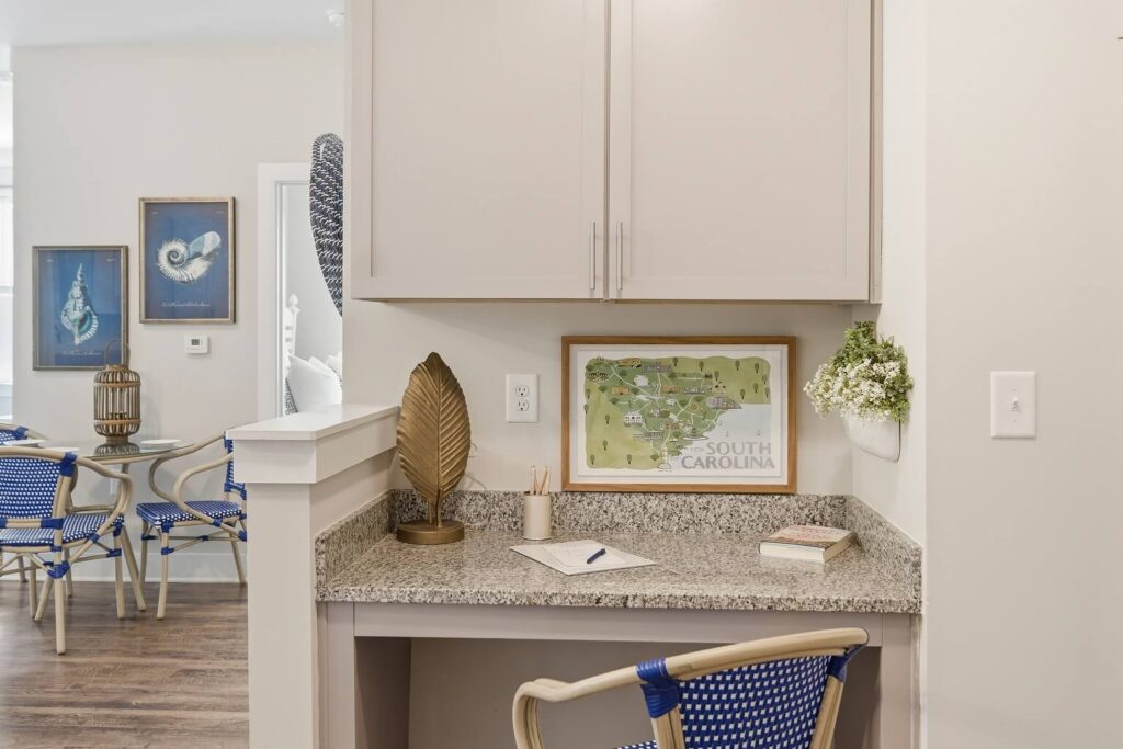 A small home office nook with a granite desk, blue chair, pencils, a notebook, decorative leaf, map art labeled South Carolina, and flowers. Blue chairs and framed art are visible in the adjoining room.