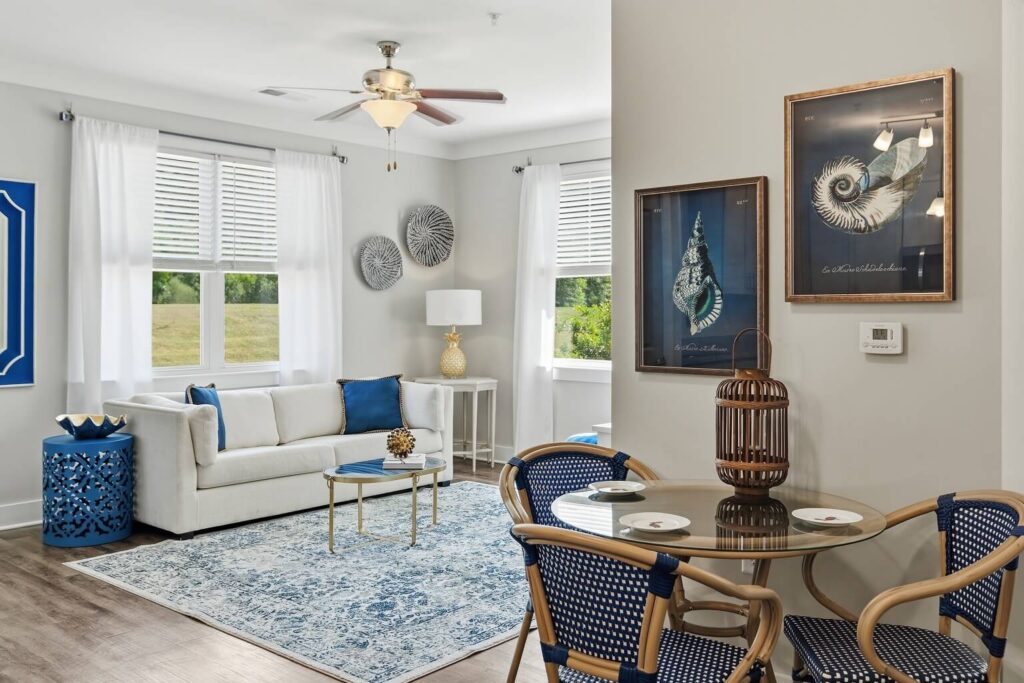 Bright living and dining area with white sofa, blue accents, round glass table, two chairs, framed seashell art, large windows with white curtains, and a blue patterned rug on light wood flooring.
