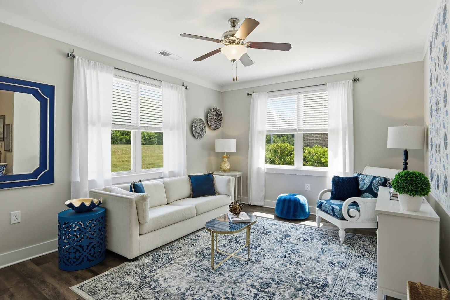 Bright living room with white walls, two couches, blue and white decor, a patterned rug, a ceiling fan, a window view of greenery, and various decorative items including pillows, lamps, and wall art.
