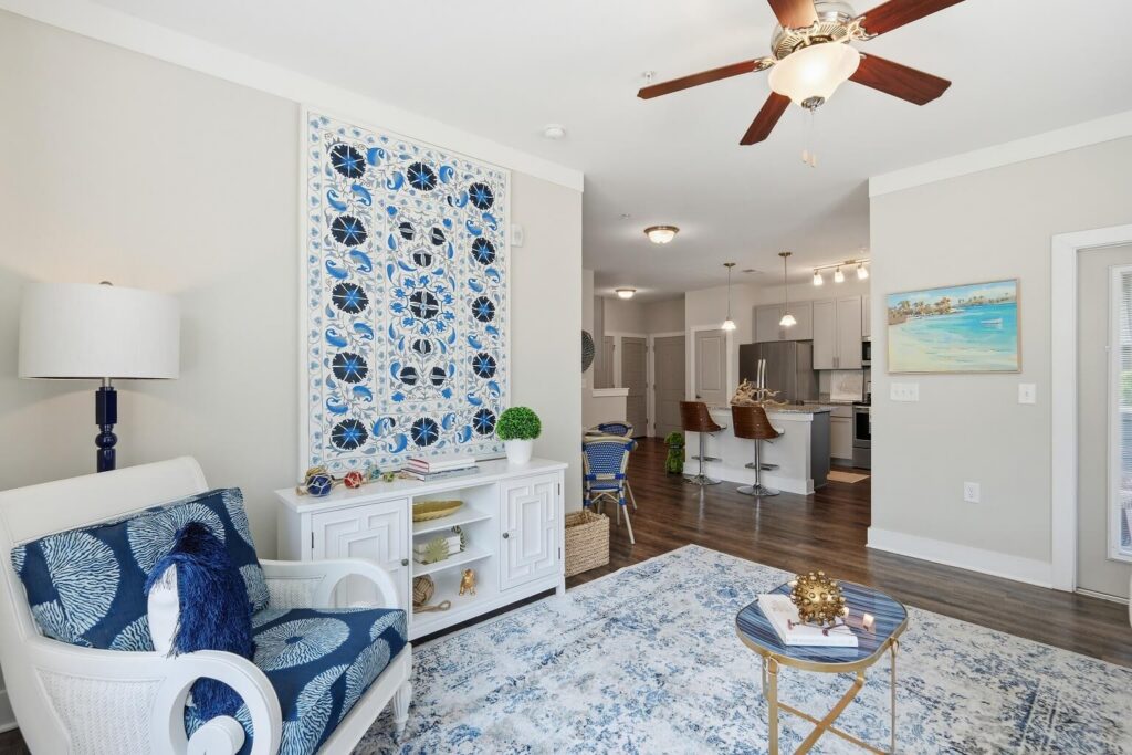 Bright living room with blue and white decor, a patterned wall hanging, an armchair, a rug, a small table, and a ceiling fan, opening to a modern kitchen with barstools and pendant lights.