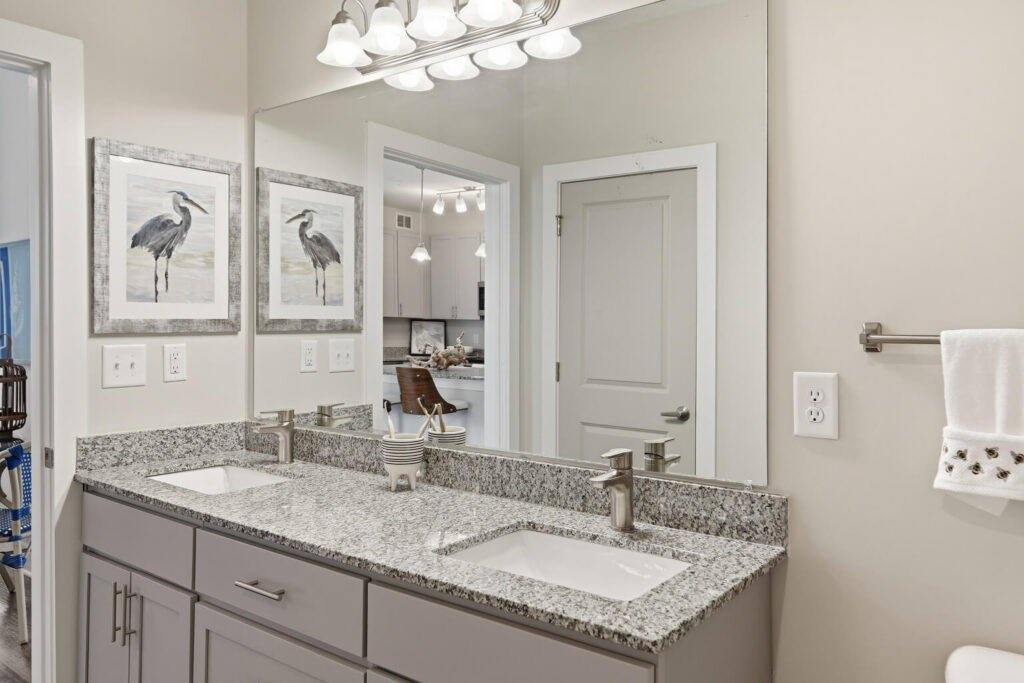 Modern bathroom with a double sink vanity, granite countertop, large mirror, and two framed heron art pieces. Reflected in the mirror is a view of a kitchen and a dining area.