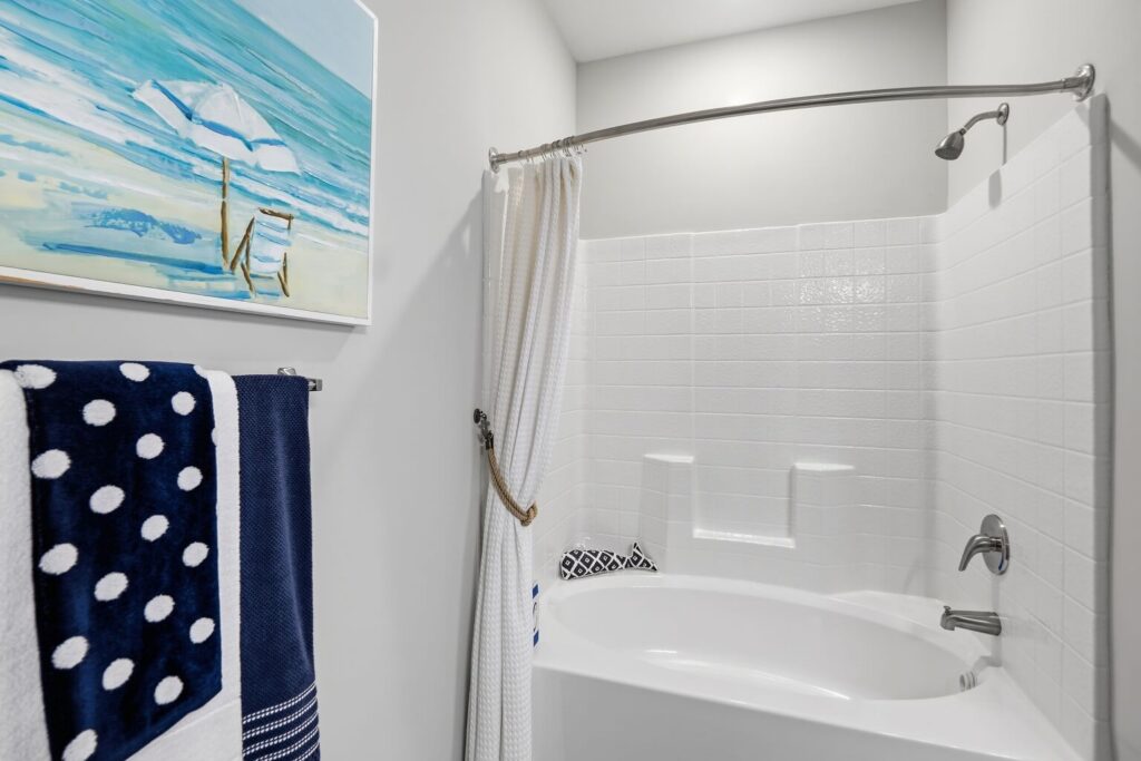 A white bathtub with a curved shower rod and white curtain, white tile surround, and navy towels with white polka dots. A beach-themed painting hangs on the light gray wall above the towels.