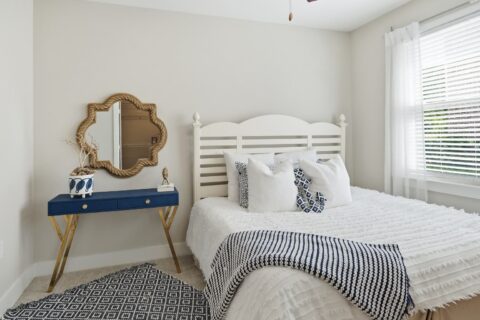 Bright bedroom with a white bed, textured bedding, navy and white pillows, a blue and white rug, a navy desk with gold legs, decorative items, a mirror, and a large window with sheer curtains.