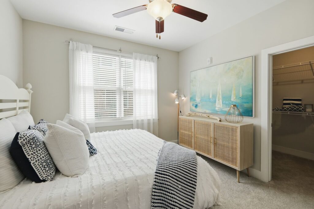 Bright bedroom with a white bed, patterned pillows, woven cabinet, large sailboat painting, floor lamp, ceiling fan, and window with sheer curtains. Open door shows a closet with shelving.