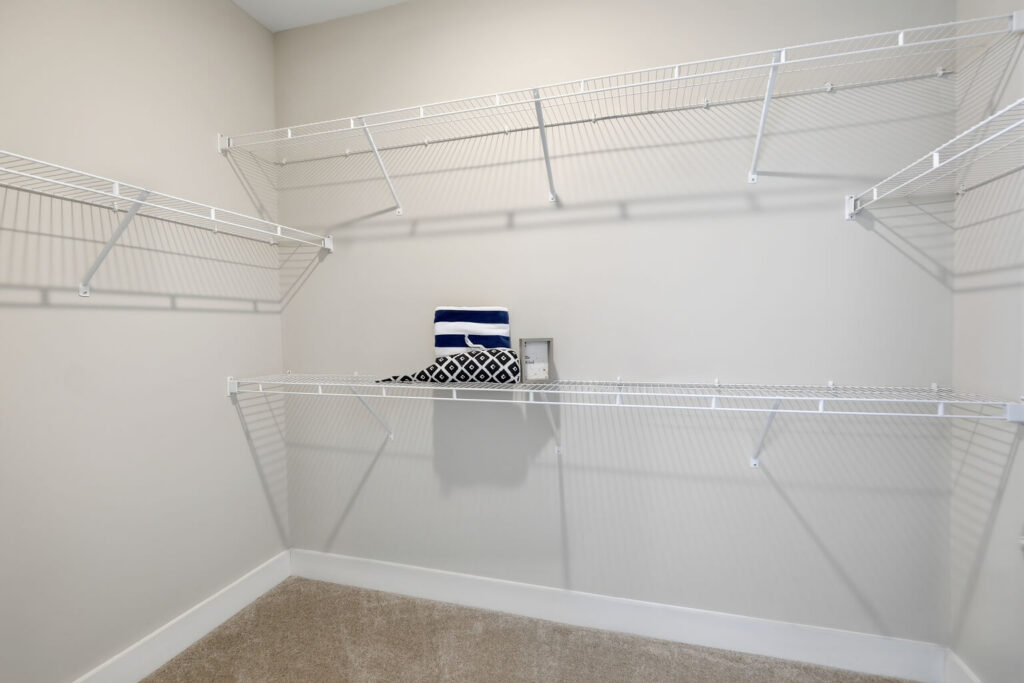 An empty walk-in closet with white wire shelves on three walls. A small stack of folded towels and a decorative pillow are placed on the middle shelf. The floor is carpeted.