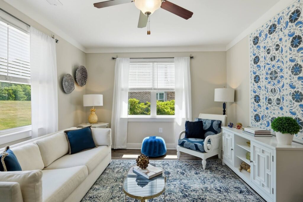 Bright living room with white sofa, blue accents, patterned rug, glass coffee table, and large blue-and-white wall art. Sunlight streams in through two windows with sheer curtains. Modern, cozy decor.