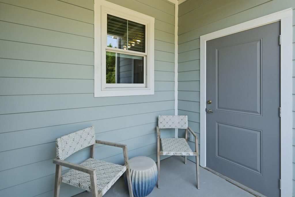 Two woven chairs and a small blue-and-white ceramic stool sit on a porch with light blue siding, a gray door, and a white-framed window.