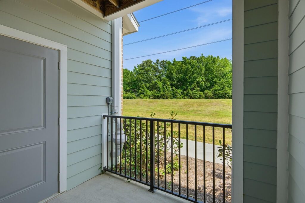 A small balcony with a gray door, black railing, and view of green grass, bushes, and trees under a clear blue sky.