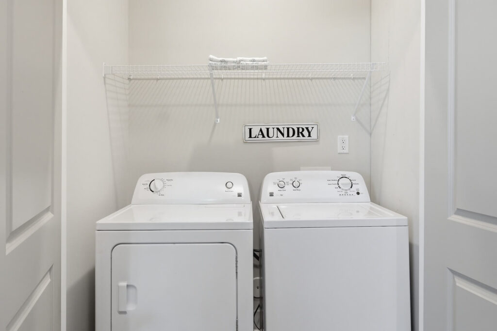 A laundry area with a white dryer and washing machine, a wire shelf holding folded towels above, and a Laundry sign on the wall. The space is bright with white walls and doors.