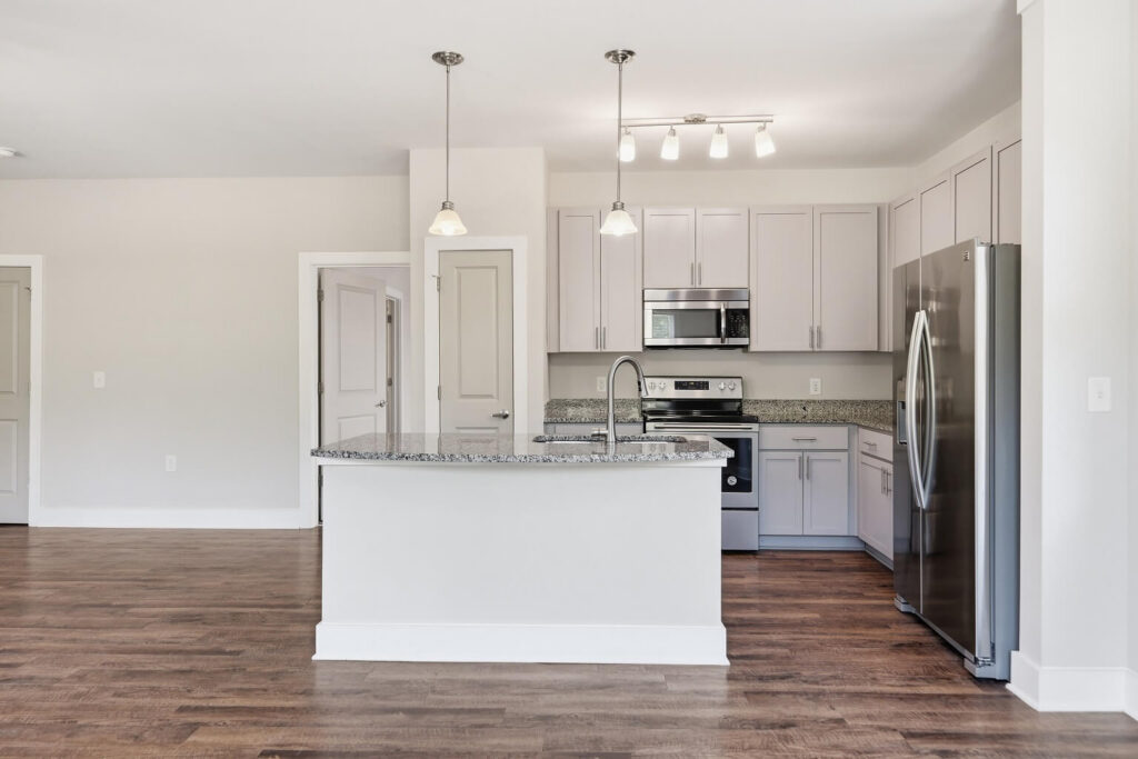 Modern kitchen with white cabinets, stainless steel appliances, granite countertops, an island with a sink, pendant lights, and wood flooring. Walls are light-colored and the kitchen is bright and spacious.