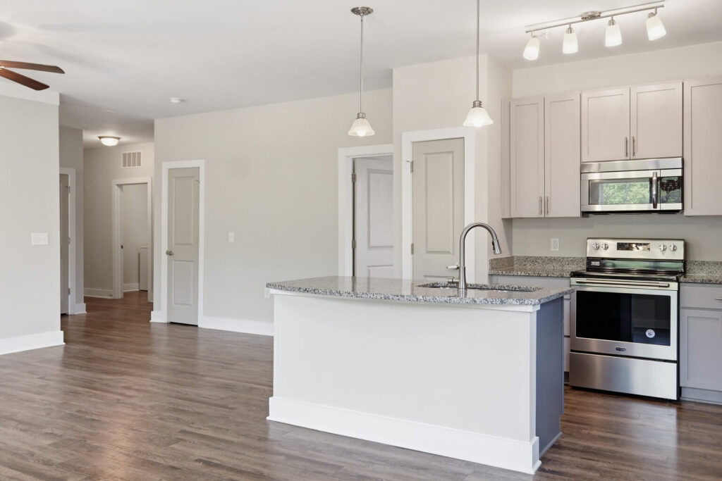 Modern kitchen with light gray cabinets, stainless steel appliances, a granite island with a sink, pendant lighting, and hardwood floors. Open layout with white walls and multiple doorways in the background.