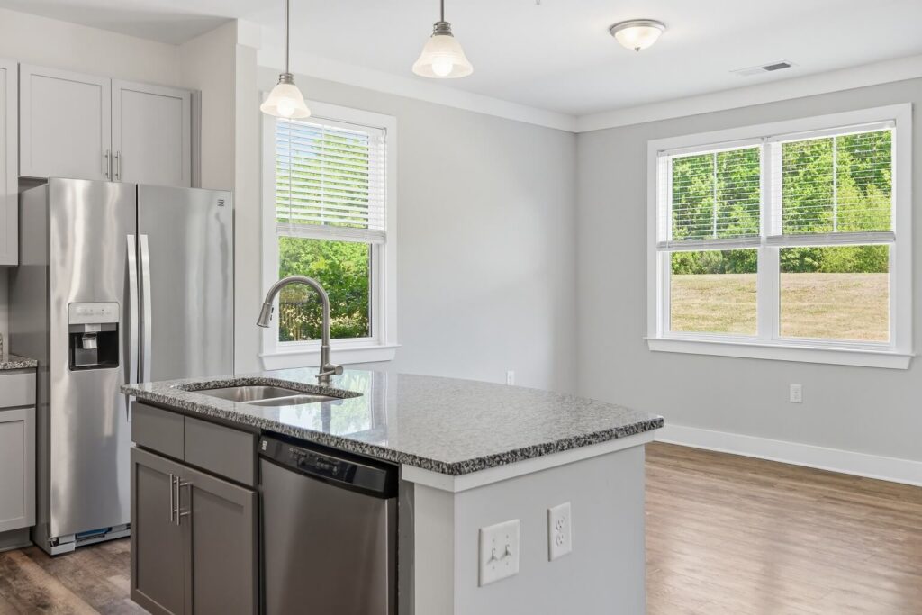 Modern kitchen with a stainless steel fridge, dishwasher, sink in a granite island, and large windows letting in natural light with a grassy view outside.